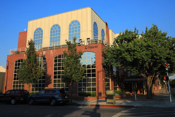 A brick building with large windows on a tree-lined street corner on a sunny day. The word Palantir appears in black lettering against the red brick.