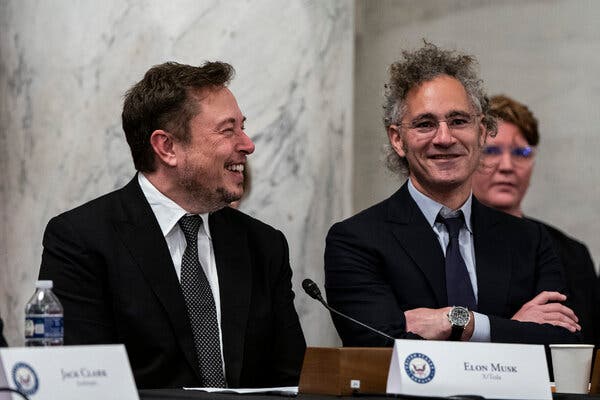 In a Senate room, Elon Musk smiles at Mr. Karp. They are seated next to each other and wearing jackets and ties.